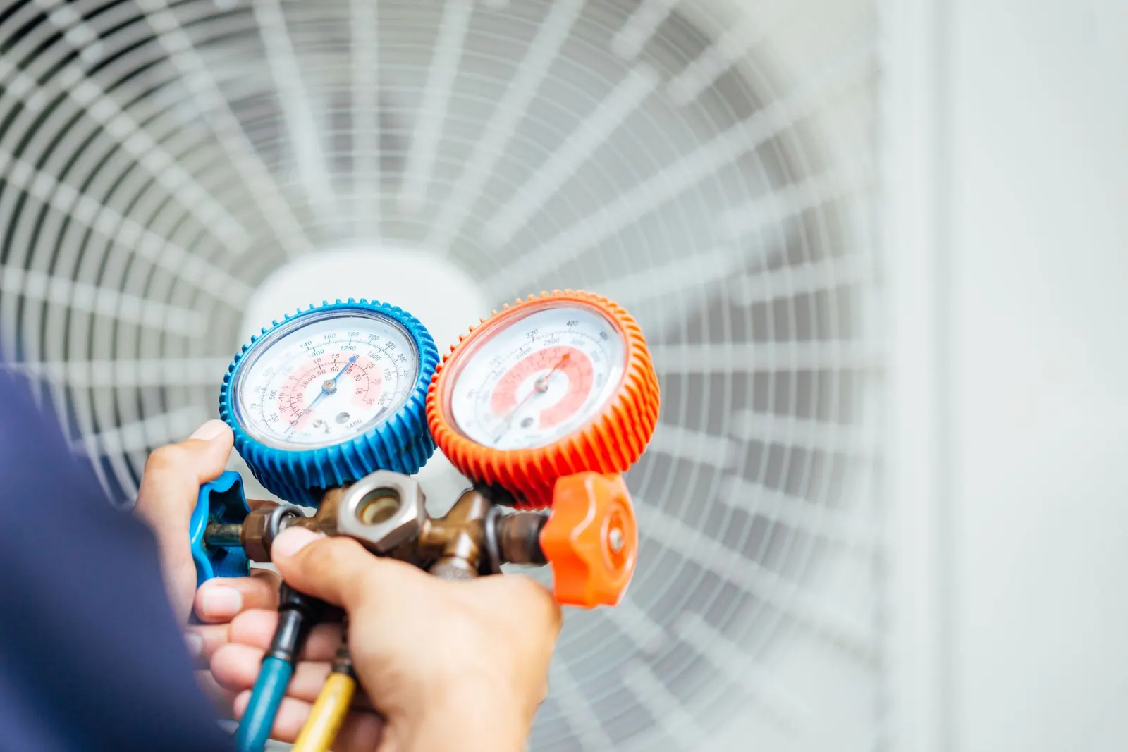 Hands holding a HVAC manifold gauge set in front of an outdoor air conditioning unit.