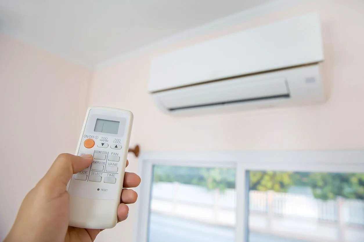 A hand holds a white remote control, pointing it toward a white wall-mounted air conditioning unit in a room.